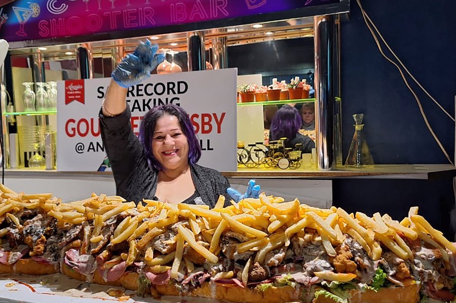 In this photo provided by Joe Redelinghuys. left, for Annies Ladies Bar, Annie Redelinghuys poses with a giant Gatsby sandwich on display at the establishment in Cape Town, South Africa, Saturday, June 29, 2024. A South African restaurant and bar has served up a 3-meter-long (9.8 feet) sandwich in the latest offering from an establishment with a taste for the supersize. (Annies Ladies Bar via AP)