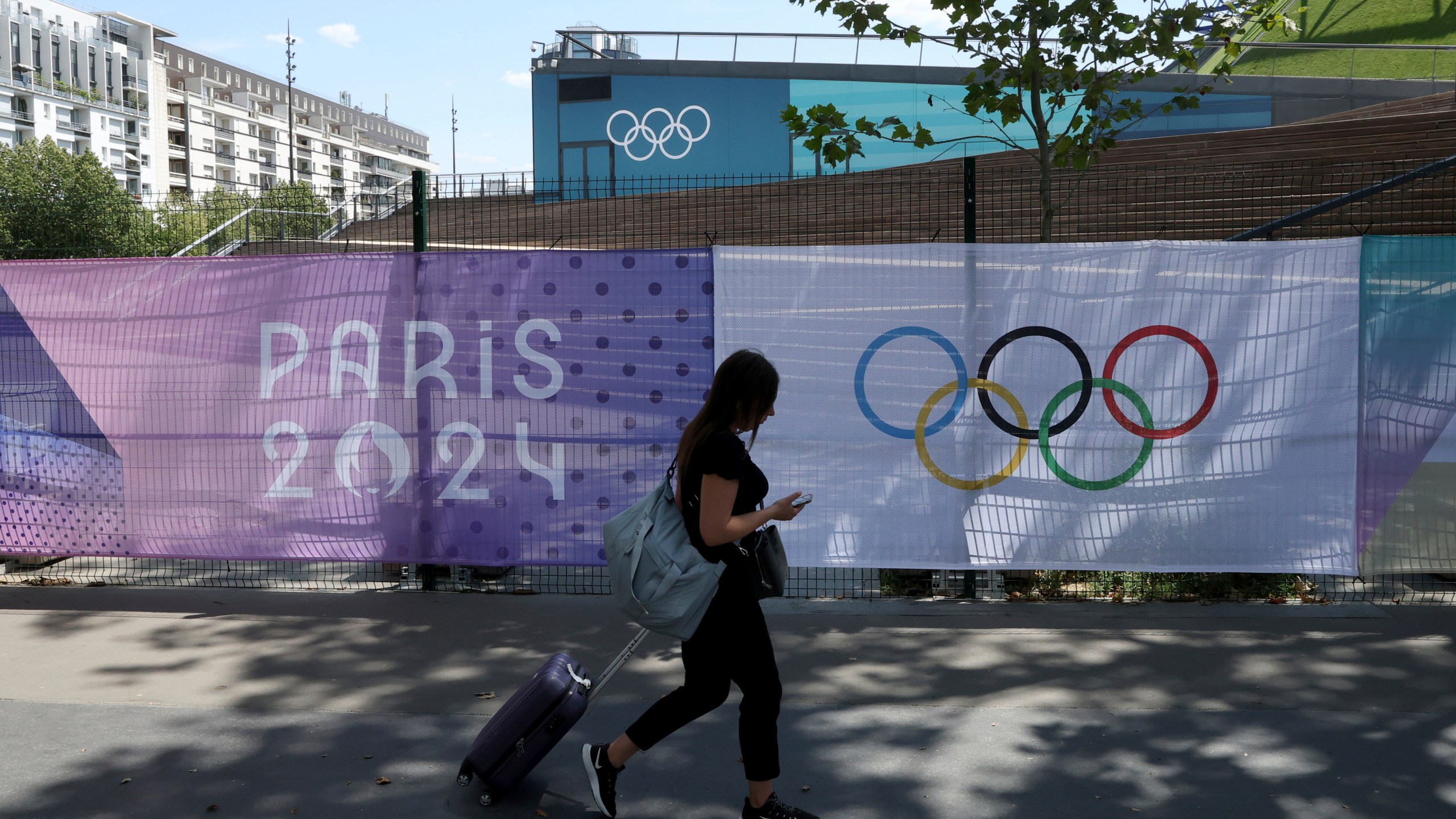 A woman pulls her luggage by Paris 2024 Olympic banners, just nine days before the start of the Paris Olympic games, Wednesday, July 17, 2024 in Paris. (AP Photo/Aurelien Morissard)